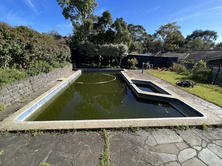 Neglected old swimming pool with stained water and overgrown backyard surrounds